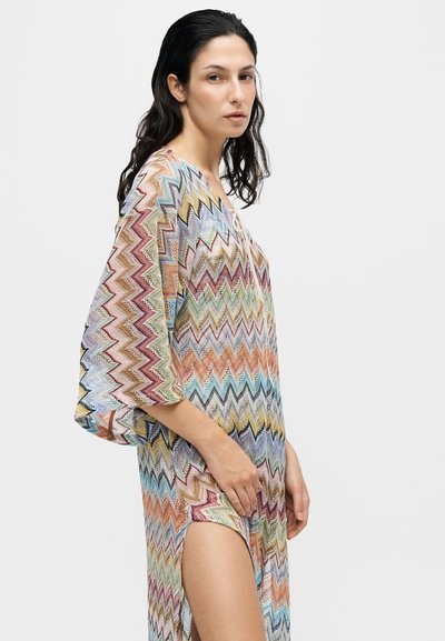 Femme aux longs cheveux foncés porte une robe colorée à motif zigzag avec de larges manches et une fente latérale haute, debout devant un fond clair uni.
