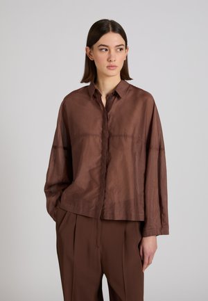 Młoda kobieta o prostych brązowych włosach, ubrana w luźną brązową koszulę zapinaną na guziki i pasujące spodnie, stoi z jedną ręką w kieszeni, spoglądając w prawo.