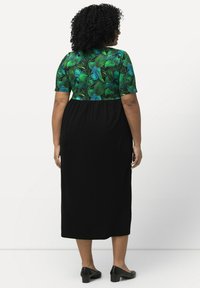 Vestido maxi negro con una parte superior de patrón de hojas verdes y mangas cortas, con una cintura ajustada y textura suave. Zapatos negros de tacón.