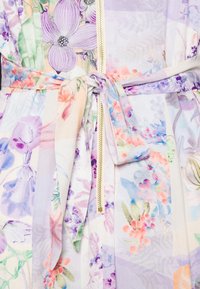 Blommönstrad tyg i pastellnyanser knuten med en rosett runt en guldlås på ett plagg.