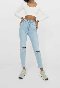Helles blaues High-Rise Skinny-Jeans mit Rissen an den Knien, kombiniert mit einem weißen, gerippten Cropped Top und neutralen Sneakers.
