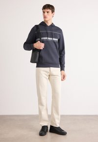 Hanorac bleumarin cu logo texturat și dungi gri, pantaloni crem și adidași negri. Bărbatul ține o geantă mică neagră. Material neted, croială lejeră.