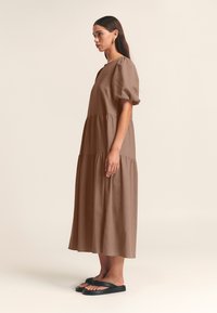 Brun midi-kjole i linned med korte pufærmer, lagdelt design og firkantet udskæring, kombineret med sorte flip-flop-sandaler.