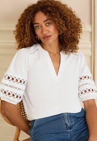 Witte blouse met een V-hals en pofmouwen. Heeft bloemige kantaccenten op de mouwen, een gladde textuur en een losse pasvorm. Gecombineerd met denim jeans.