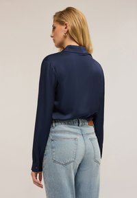 Donna con capelli biondi che indossa orecchini a cerchio dorati, una blusa blu navy a maniche lunghe e jeans a vita alta azzurri, di spalle.