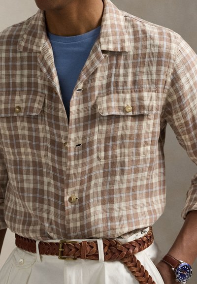 Καφέ και εκρού πουκάμισο με καρώ μοτίβο και κοντά μανίκια, με δύο τσέπες στο στήθος, συνδυασμένο με μπλε T-shirt με στρογγυλή λαιμόκοψη. Φαίνονται ζώνη πλεκτή σε ταμπά απόχρωση και ρολόι.