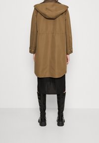 Cappotto impermeabile marrone con cappuccio, polsini a bottone a pressione e vita cinctata; indossato sopra un vestito nero e stivali neri fino al ginocchio.