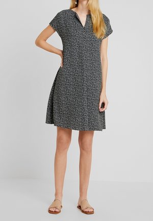 Schwarzes Kleid mit weißem Blumenmuster, kurzen Ärmeln, V-Ausschnitt und ausgestelltem Design. Kombiniert mit beigen flachen Sandalen.