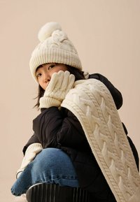 Krēmskrāsas adīta cepure ar pom-pomu, atbilstošas cimdus un adītu šalli ar kabeli, apvienota ar melnu pūkaini jaku un ziliem džinsiem.