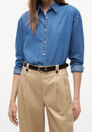 Personne portant une chemise en jean bleue à boutons avec manches retroussées, rentrée dans un pantalon beige à plis taille haute avec une ceinture noire.