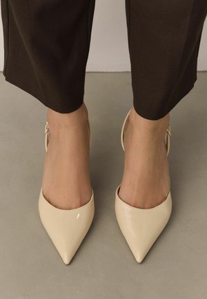 Woman wearing beige patent leather pointed-toe slingback heels with brown cropped trousers standing on a gray floor.