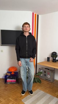 Svart hettejakke med glidelås og frontlommer, kombinert med lyseblå jeans og svarte sneakers. Enkelt design og avslappet passform, med en teksturert bakgrunn.