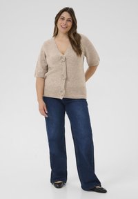Cardigan en tricot beige avec un col en V, des manches courtes et trois boutons floraux. Associé à un jean flare bleu foncé et des chaussures plates noires.