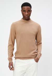 Suéter de punto beige con un patrón texturizado, cuello alto y puños ajustados, combinado con jeans blancos. Tejido suave con detalles acanalados.