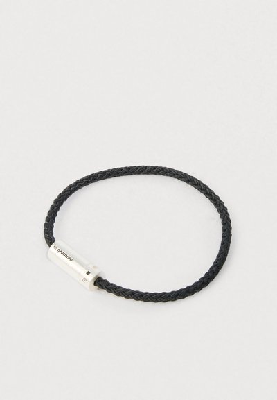Pulseira de cordão trançado preto com um fecho cilíndrico prateado gravado com "le gramme" e "7g" sobre um fundo branco.