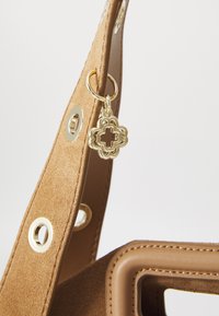 Brun ruskind yndlingstaske rem med guldtone detaljer og dekorativt vedhæng med blomstermotiv. Glatte læderdetaljer og syede detaljer.