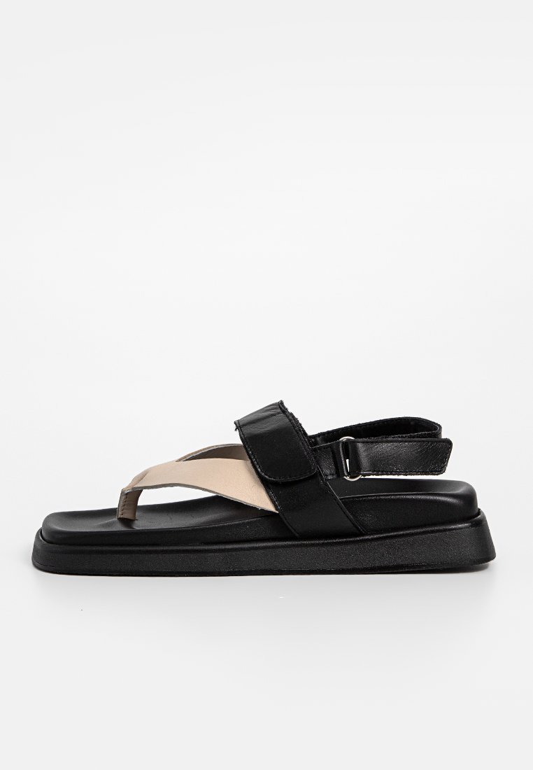 Schwarze und beige Sandale mit flacher Sohle, Gummibasis und einem Riemen mit verstellbarem Schnallenverschluss. Das Material wirkt synthetisch und glatt.