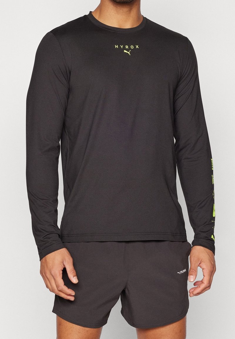 Černé sportovní tričko s dlouhým rukávem s reflexními žlutými nápisy "HYROX" a detaily na rukávech. Jemná, lehká látka. Klasický střih.