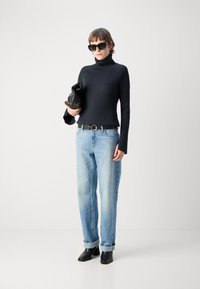 Maglione nero a costine con colletto alto, jeans dritti blu chiaro con orli arrotolati e stivali neri in pelle alla caviglia. Tiene una borsa a mano nera.