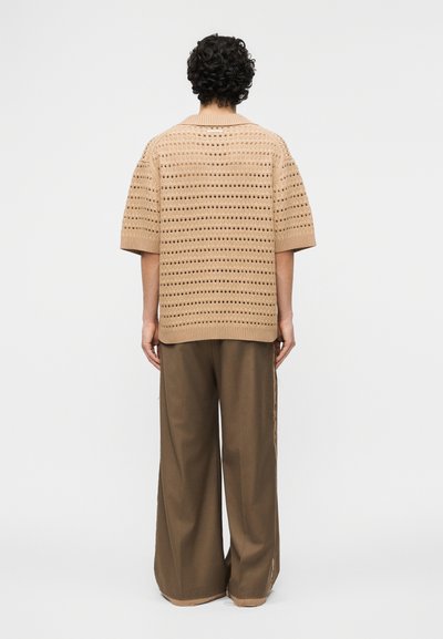 Άτομο με μαλλιά σε μπούκλες στέκεται πλάτη, φοράει μπεζ πουλόβερ με ανοιχτή πλέξη και κοντά μανίκια και καφέ φαρδιά παντελόνια.