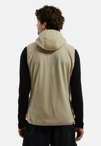 Lightweight beige vest with a hood, featuring a textured pattern and a fitted design. Long black sleeves visible underneath.