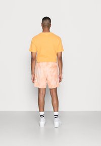 Camiseta de algodón naranja para hombre, combinada con pantalones cortos de tie-dye naranja claro. Zapatillas blancas y calcetines a rayas negras completan el conjunto.