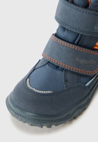 Sapatilha infantil azul escuro com um design em camurça e malha, fechos em Velcro, e uma sola de borracha texturizada com pormenores em laranja.