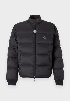 Homme portant une doudoune noire, un sweat zippé noir avec un motif blanc, un jean bleu clair et des baskets blanches, debout avec la main dans la poche.