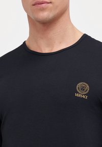 Zwarte katoenen t-shirt met een ronde halslijn. Kenmerkt zich door een gouden geborduurd Versace-logo en hoofdembleem op de linkerborst.