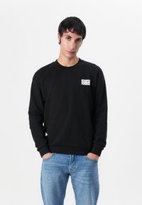 Felpa nera in cotone con maniche lunghe e polsini a costine. Presenta una piccola etichetta con logo bianco sul lato sinistro del petto. Abbinata a jeans azzurri.