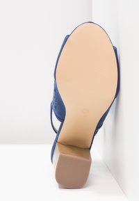 Chaussure à talon haut en daim bleu avec une semelle en caoutchouc texturée, présentant des découpes sur le côté et un talon empilé robuste.