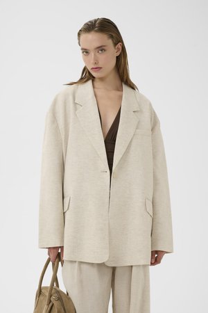 Frau trägt einen übergroßen hellbeigen Blazer und passende Hose, hält eine beige Handtasche, mit braunem tiefem V-Ausschnitt darunter.