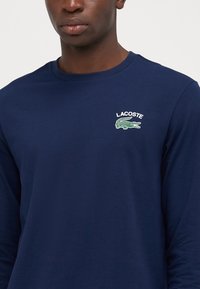 Chemise à manches longues bleu marine en coton, avec un logo crocodile brodé vert et le texte "LACOSTE" sur la poitrine gauche. Texture lisse.