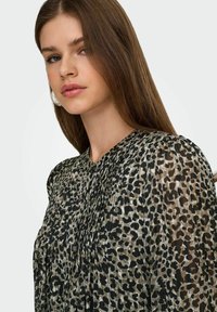 Blouse transparente à motif léopard noir et beige, avec des plis au niveau de l'encolure et un tissu doux et fluide.