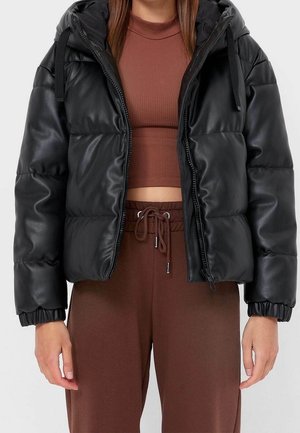 Femme portant une veste matelassée noire à capuche par-dessus un haut court côtelé marron et un pantalon marron à cordon de serrage, debout avec les bras détendus.