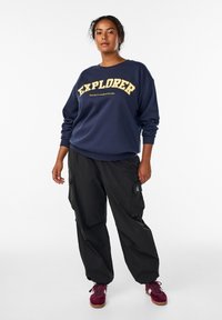 Jūrininkų megztinis su auksiniu užrašu „EXPLORER“; juodos krovinių kelnės; bordo spalvos sportbačiai; laisvas kirpimas; minkšta medžiaga; raištinės rankovės ir apačia.