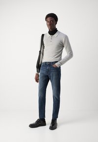 Jasnoszary t-shirt henley z trzema guzikami, w połączeniu z ciemnoniebieskimi dżinsami i czarnymi skórzanymi butami. Model nosi czarną torbę na ramię.