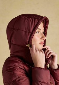 Burgundy puffer jacket with a hood, featuring a quilted design, shiny texture, and gold hardware accents. Model adjusts the hood.