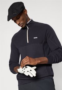 Pullover preto com fecho 1/4, detalhes em bege, tecido texturizado, logo "BOSS" em branco. Modelo usa luva de golfe branca, boné preto.