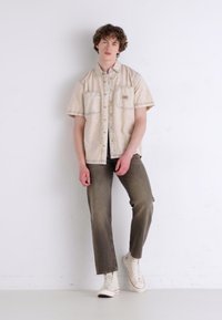 Kortermet beige skjorte med frontlommer, detaljert søm, kombinert med løse olivengrønne bukser og hvite høye sneakers.