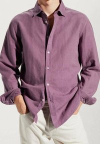 Camisa de lino morada con cuello abotonado, mangas largas remangadas en los puños y un corte holgado. Llevada sobre pantalones de color claro.