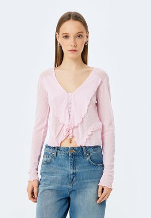 Lys pink cropped cardigan med flæser foran, fem knaplukninger og lange ærmer. Båret over højtkøbte blå denim jeans.