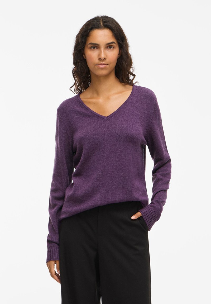 Maglione viola a V con maniche lunghe, polsini a coste e tessuto morbido, abbinato a pantaloni neri larghi. Design semplice con dettagli minimi.