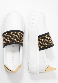 Białe sneakersy wsuwane z czarnym elastycznym paskiem ozdobionym wzorem z złotych łańcuchów. Złote akcenty na zaokrąglonym nosku i pięcie. Teksturowana podeszwa.