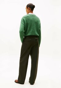 Donna con i capelli raccolti in un bun, che indossa un maglione verde, pantaloni scuri a righe con tasche posteriori con bottoni e scarpe marroni, in piedi con le mani nelle tasche.