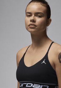 Sosten negro deportivo con tirantes finos, que presenta un logo Jumpman blanco en el pecho y una banda contrastante con la inscripción "Jordan". Textura suave.