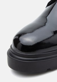 Biqueira de bota em couro patente preto brilhante com sola de borracha preta grossa, em close-up mostrando a textura lisa e brilhante e a parte frontal arredondada.
