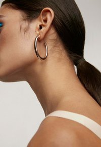 Pendiente de aro de oro rosa con una superficie lisa y reflectante, sujeto a una oreja. El cabello está recogido hacia atrás, mostrando el diseño del pendiente.