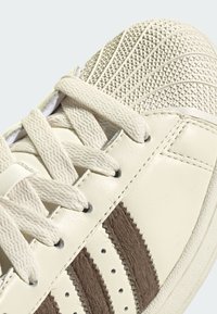 Biela kožená teniska s textúrovanou, svetlohnedou prstovou časťou a hnedými chlpatými pruhmi. Má ploché šnúrky a malé strieborné očká.