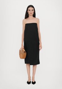 Svart off-shoulder kjole med rett snitt og knelengde. Modellen holder en vevd beige håndveske. Sorte spisse flate sko fullfører antrekket.